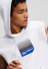 Felpa senza maniche bianca con cappuccio, caratterizzata da un design grafico con strisce orizzontali nere e blu e il testo "KARL LAGERFELD JEANS".