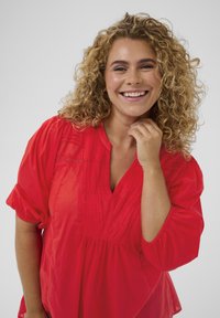 Blouse rouge avec un décolleté en V, des manches bouffantes courtes et des plis verticaux. Texture lisse avec une coupe légèrement ample et des détails minimaux.