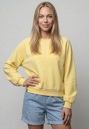 Blonde Frau steht mit einer Hand in der Hüfte, trägt ein gelbes Sweatshirt und hellblaue Jeansshorts vor einem einfarbigen grauen Hintergrund.