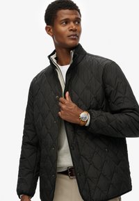 Sort quiltet jakke med høj krave, med trykknapper og et subtilt logo. Bæres over en lys grå sweater, parret med et ur.
