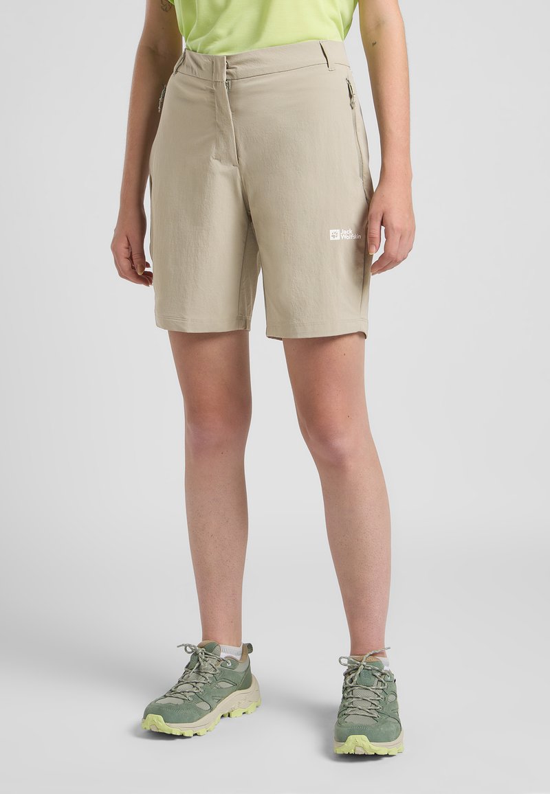 Beige Shorts verfügen über eine Reißverschlusstasche an der Seite, einen flachen Frontbund und einen leichten Stoff. Kombiniert mit grünen Wanderschuhen für den Außeneinsatz.