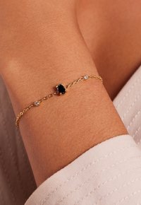 Gouden kettingarmband met een ovale zwarte edelsteen en twee kleine heldere accenten, met een verfijnd ontwerp en een gladde textuur.