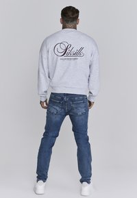 Grauer Sweatshirt mit bedrucktem Logo auf der Rückseite, kombiniert mit blauen Jeans und weißen Sneakers. Lässiger Schnitt, weicher Stoff und gerippte Bündchen.