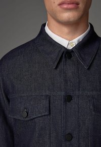 Denim jaak struktuurse kraega, tume indigo värvi, mustad nööbid ja kaks rinna taskut. Valge särk väljub krae alt.