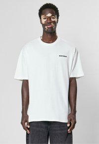 Tricou din bumbac alb, supradimensionat, cu mâneci scurte, guler rotund și text negru "9N1M SENSE." pe partea stângă. Purtați cu blugi închiși la culoare.
