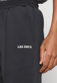 Čierne športové nohavice s hladkou textúrou, s bielym logom "LES DEUX" na stehne. Elastický pás a bočné vrecká sú viditeľné.