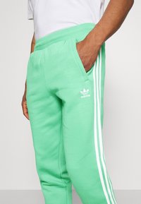 Muž v jasně zelených teplácích Adidas s bílými pruhy a logem, ruka v kapse, v kombinaci s bílým tričkem.