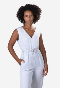 Jumpsuit sans manches bleu clair avec un col en V, taille cintrée et une ceinture blanche assortie. Texture de tissu lisse, poches sur les côtés.