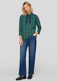 Karierte Bluse in Blau und Grün mit dreiviertel Ärmeln, rundem Ausschnitt und marineblauen Akzentbändern, kombiniert mit weiten Jeanshosen.