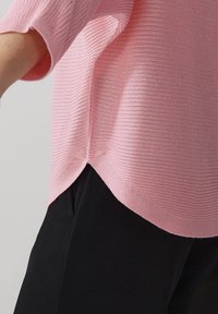 Haut en tricot rose avec une texture côtelée, manches courtes et ourlet incurvé, associé à un pantalon en tissu noir. Vue rapprochée des détails du tissu.
