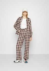 Conjunto de camisa e calças xadrez em castanho e creme, combinado com uma blusa branca e um boné branco. Os sapatos apresentam um design multicolorido.