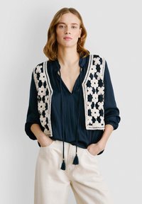 Mornarska bluza s bijelim cvjetnim kukičanim prslukom, koji ima nabran podstavljanje i vezivanje. Hlače su svijetlo bež boje, s bočnim džepovima.