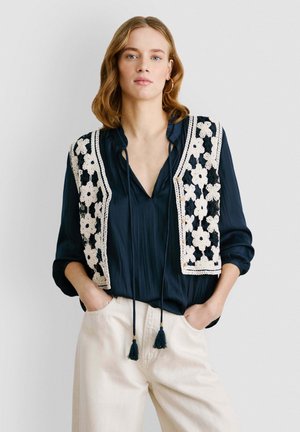 Blusa marina con un chaleco de flores blancas de ganchillo, que presenta un cuello con volantes y cierre de lazo. Los pantalones son de un beige claro, con bolsillos laterales.