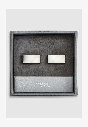 St orter i rustfrit stål i rektangulær form med en børstet overflade, præsenteret i en grå æske beklædt med sort ruskind.