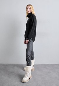 Kvinde med langt blont hår står mod en grå væg iført sort turtleneck-sweater, grå stribede bukser og hvide støvler med pelsfor.