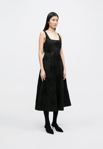 Μαύρο αμάνικο φόρεμα με με floral κεντήματα από χάντρες, τετράγωνη λαιμόκοψη, ρέουσα φούστα και διακριτική γυαλάδα, συνδυασμένο με μαύρους καλσόν και παπούτσια.