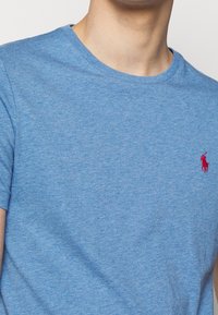 Blue cotton t-shirt with a crew neck and short sleeves. Features a small red embroidered logo on the chest. Soft texture.