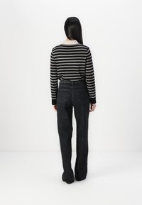 Kvinne med langt svart hår iført en svart og hvit stripete langermet skjorte og høytlivde svarte jeans som står mot en hvit vegg.