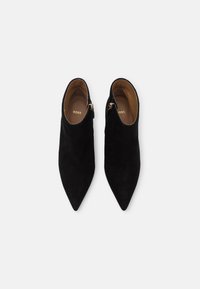 Botines de ante negros con puntas afiladas y cremalleras laterales. Textura suave con un interior beis y una marca dorada visible en el interior.
