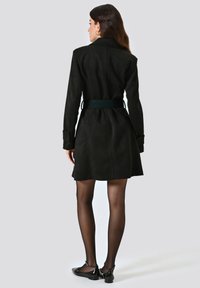 Cappotto in suede nero con cintura, maniche lunghe, vestibilità sagomata e lunghezza al metà coscia, caratterizzato da una finitura liscia e sottili dettagli testurizzati.