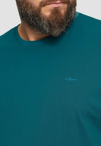 Turkusowa koszulka z okrągłym dekoltem, wykonana z miękkiej bawełny, z małym niebieskim logo "s.Oliver" na piersi. Gładka tekstura, luźny krój.