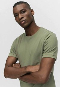 Olivgrön, figursydd t-shirt med korta ärmar, rund halsringning och ribbat detaljer vid ärmslå enden, bärs av en person med armarna i kors.