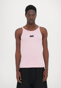 Lys rosa ribbet tanktop med smale stropper og rund hals, med et lite svart logo merke foran. Brukt med svarte bukser.