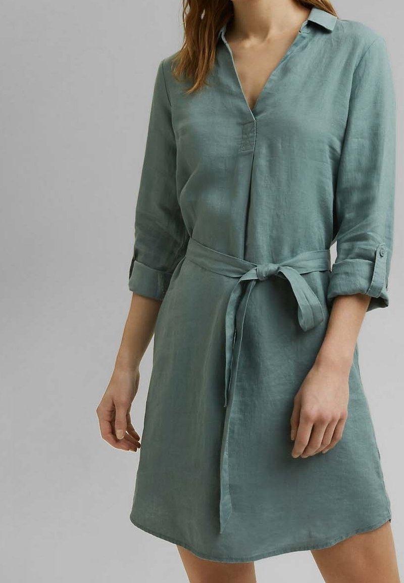 Robe-chemise en lin vert clair avec col, manches longues retroussées et taille ceinturée. Elle présente une patte de boutonnage à l'avant et un ourlet arrondi.