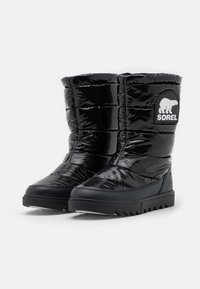 Stivali isolati neri con finitura lucida, caratterizzati da un design imbottito, suola in gomma e una toppa circolare con un grafica di un orso polare.