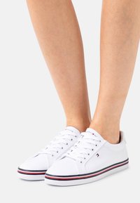 Sneakers bianchi in tela con punte tonde, lacci tradizionali e strisce blu navy e rosse a contrasto sulla suola. Suola in gomma per una buona aderenza.