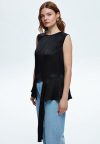 Top nero senza maniche con pois testurizzati, orlo svasato e un lungo fiocco decorativo. Abbinato a jeans a vita alta di colore blu chiaro.