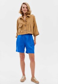 Kort bruin linnen jasje met twee voorzakken, gecombineerd met felblauwe shorts en beige sneakers. Het jasje heeft grote knopen.