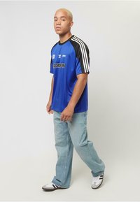 Blå atletisk skjorta med svarta ärmar, prydd med vita Adidas-ränder och logotyp, ihop med ljusa jeans och sportskor.