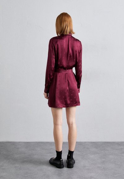 Vestido de satén burdeos con una silueta ajustada y mangas largas, que presenta un patrón de estrellas y una cintura ceñida con cinturón, combinado con zapatos negros.
