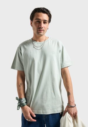 Lysegrøn bomuldst-shirt med korte ærmer, rund hals og en lille præget logo. Bruges med sølvfarvede lagdelte kæder og blå jeans.