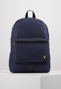 Sac à dos en tissu bleu marine avec une surface texturée, une poche zippée à l'avant et un logo d'aigle jaune en bas à droite. Poignée de transport en haut incluse.