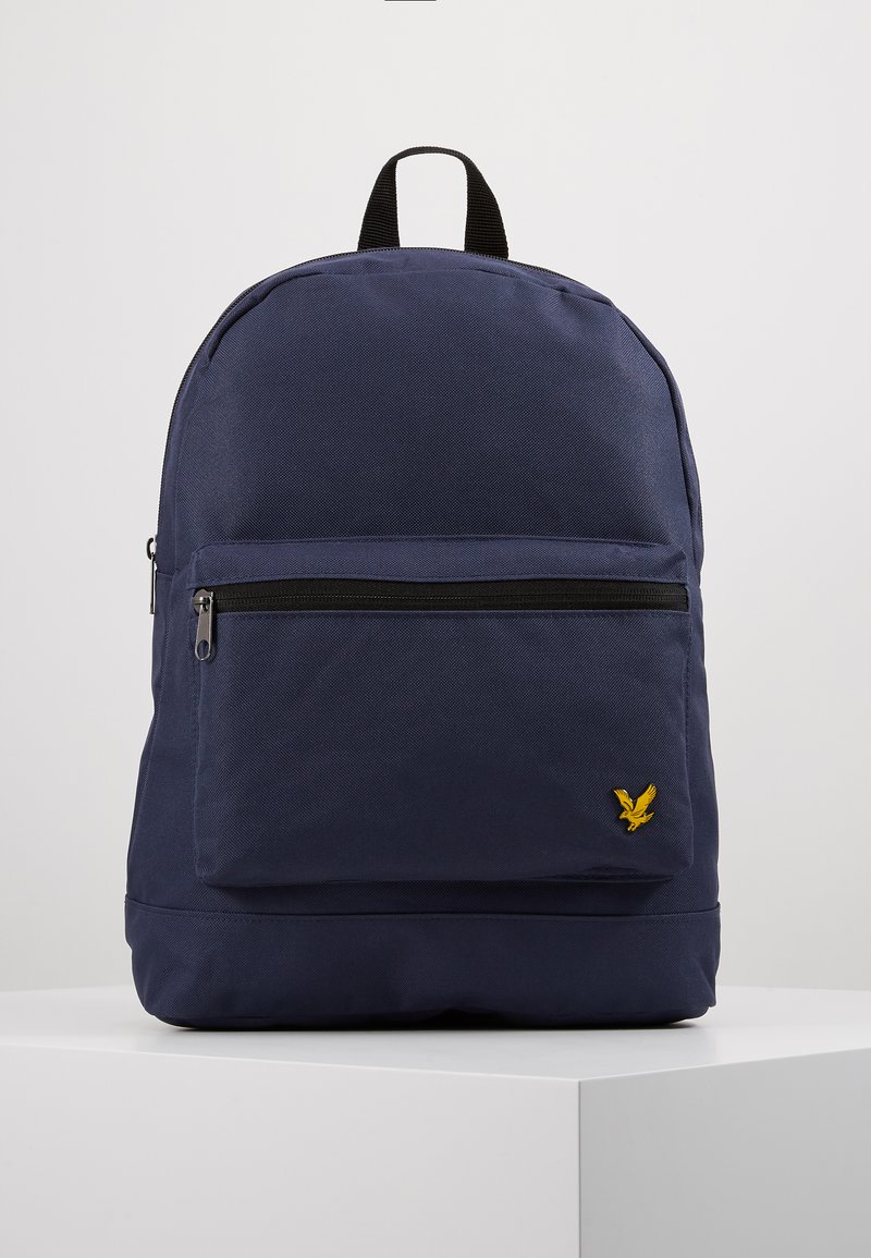 Sac à dos en tissu bleu marine avec une surface texturée, une poche zippée à l'avant et un logo d'aigle jaune en bas à droite. Poignée de transport en haut incluse.