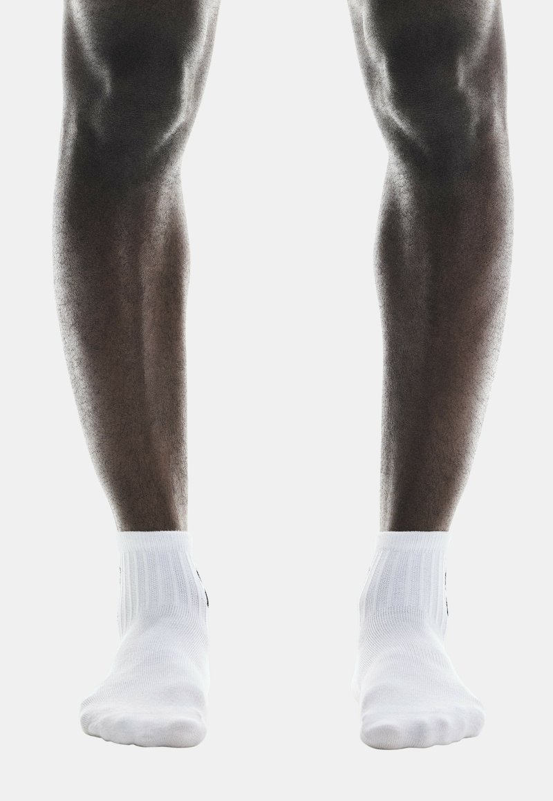 Calcetines blancos hasta el tobillo con puños acanalados se llevan en pies descalzos, con piernas lisas y musculosas visibles. El fondo es blanco liso.