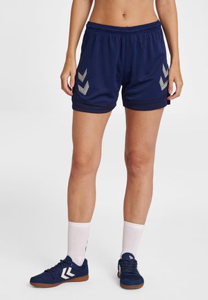 Osoba ubrana w granatowe sportowe spodenki z szarymi wzorami w jodełkę, białe skarpetki do kostek oraz granatowe buty sportowe z białymi wzorami w jodełkę.