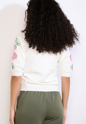 Žena s kučeravými vlasmi, oblečená v bielom krátkemu svetri s kvetinovou výšivkou na rukávoch a olivovozelených nohaviciach, zobrazená zozadu.