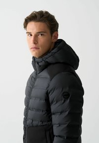 Schwarze, kapuzenlose Puffjacke mit gestepptem Design, featuring glattem Stoff, schwarzen Akzenten und einem Logo-Detail am Ärmel.