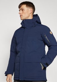 Marineblå isolert jakke med høy krage, flere lommer og sidelomme med glidelås. Har en jevn tekstur og justerbar hette.