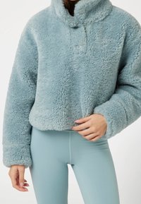 Světlě modrý pulovr z umělé kožešiny s vysokým límcem, dvěma knoflíky a krátkým střihem. Měkká, nadýchaná textura kontrastuje s hladkými, přiléhavými legínami.