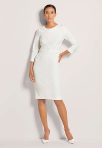 Fitted white dress with three-quarter sleeves, ribbed texture, and diagonal seam details. Paired with pointed white heels.