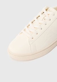 Tuvplāns krēmkrāsas sporta apaviem ar auklām un smalki izšūtu logotipu sānos, fotografēts uz gaiša fona.