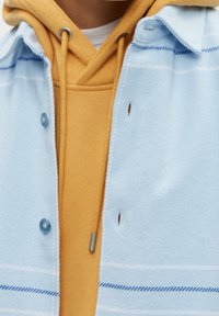 Camicia a quadri blu chiaro con bottoni blu e una felpa gialla sotto con cordino, che mostra tessuti testurizzati e dettagli di cucitura.