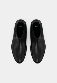 Pantofi eleganți slip-on din piele neagră cu detalii brogue și inscripția "BOSS" în interior, văzuți dintr-o vedere de sus pe un fundal alb.