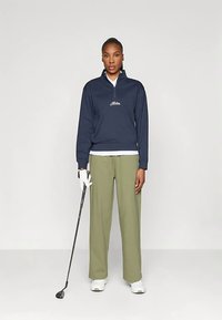 Marine blauwe fleece pullover met rits en wit logo, gecombineerd met losse olijfgroene broeken en witte sneakers. Golfclub in de rechterhand.