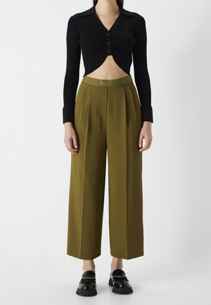 Kvinde iført olivengrønne vidde bukser med høj talje og sort cropped langærmet top med knapper, stående mod en ensfarvet hvid baggrund.