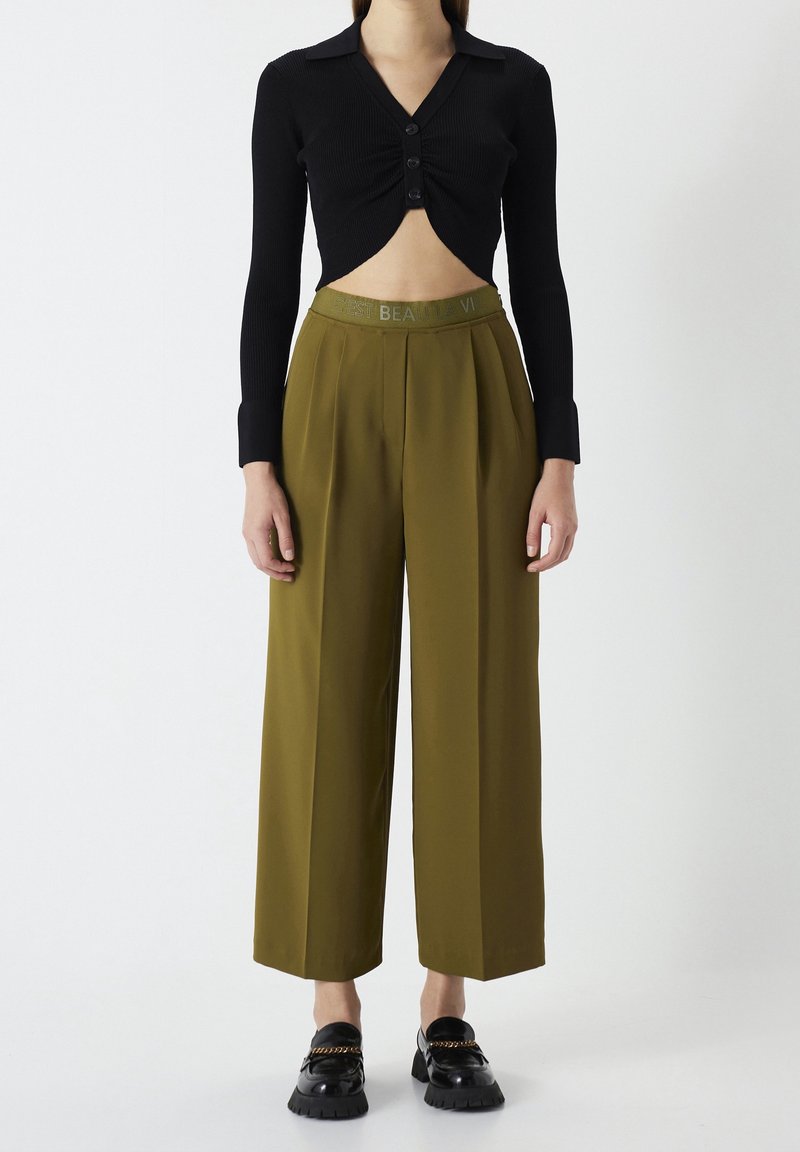 Femme portant un pantalon large olive taille haute et un top noir court à manches longues avec des boutons, debout devant un fond blanc uni.